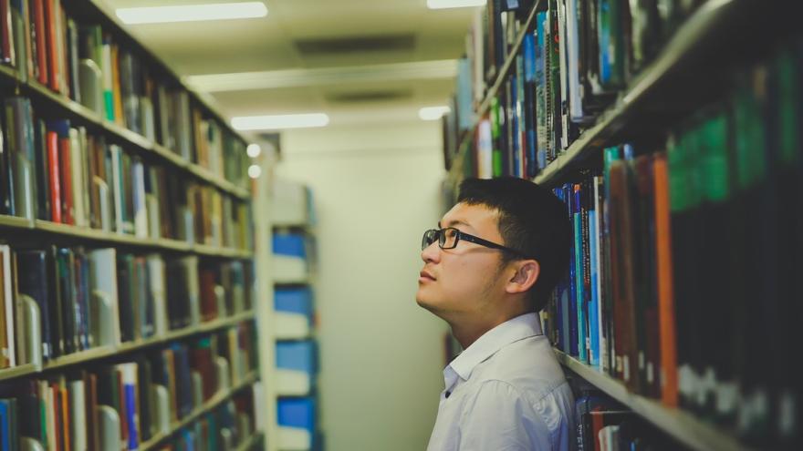 Student in einer Bibliothek, suchend vor einem Regal mit Büchern.