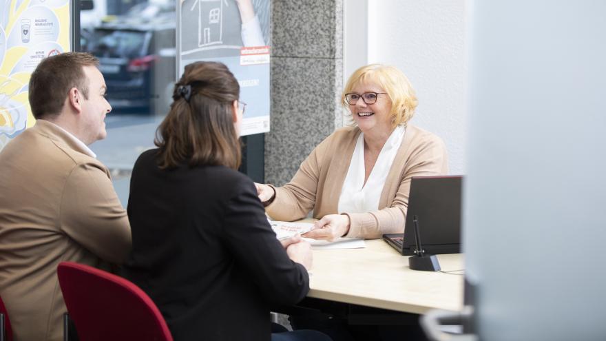 Ein Mann und eine Frau sitzen vor einem Schreibtisch. Hinter dem Schreibtisch sitzt eine lächelnde Frau. Das Bild zeigt eine Beratungssituation in einer Verbraucherberatungsstelle.