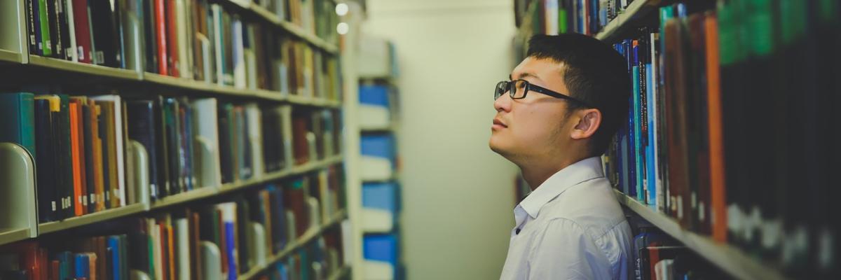 Student in einer Bibliothek, suchend vor einem Regal mit Büchern.